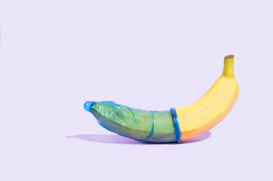 A yellow banana on a lavendar background with a blue condom rolled halfway up the banana.